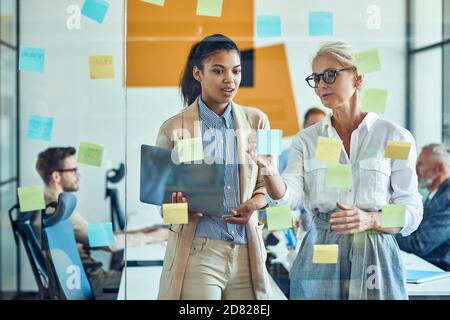 Brainstorming. Two happy female coworkers putting colorful sticky notes on a glass wall, sharing ideas and smiling while standing in the modern office Stock Photo