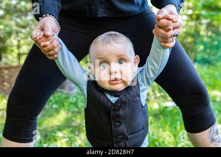Portrait of newborn baby standing with parent hand support wearing formal clothing posing for photoshoot in outdoor garden Stock Photo