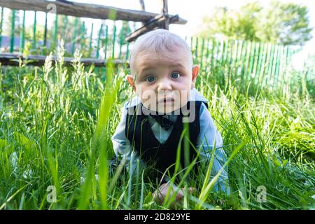 Portrait of fearful expression infant baby boy wearing formal clothing with bow sitting and lying on grass on stomach in outdoor park Stock Photo