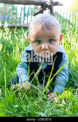 Portrait of infant baby boy, wearing formal clothing with bow sitting and lying on grass on stomach in outdoor park looking with wide open eyes Stock Photo