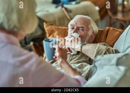 Sick elderly man lying on the couch and using his napkin for a runny nose with wife next to him. Care and health concept Stock Photo
