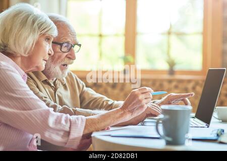 Side view of senior surprised couple pointing their fingers at laptop screen with window on the background. Copy space. Family and household concept Stock Photo