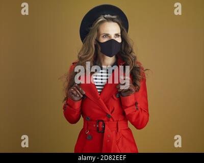 Life during coronavirus pandemic. modern woman in red coat with black mask looking into the distance against bronze background. Stock Photo