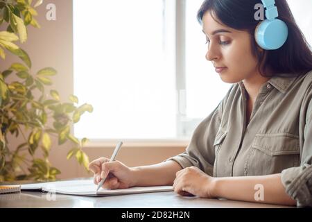 Indian woman wearing headphones writing in notebook listening course at home. Stock Photo