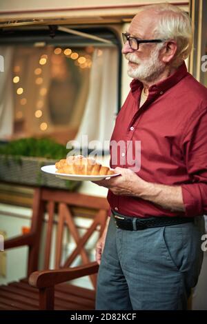 Side view of happy bearded elderly man standing near the camper van and holding a plate with croissant. Travel concept Stock Photo