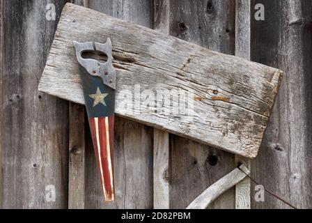 old hand saw painted with white star on blue background and red and white stripes hanging on the side of a rustic gray weathered wood barn Stock Photo