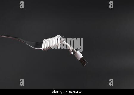 Charging mobile phone cable wrapped around fork like spaghetti. Dark grey background with fork and power cable in front. Conceptual photograph. Stock Photo
