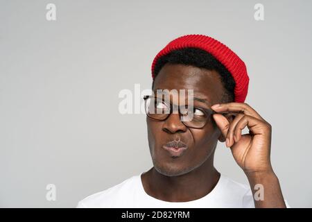 Close up studio portrait of unsure doubtful Afro American man in red hat holding eyeglasses, peers, suspicious looking away at blank copy space for ad Stock Photo