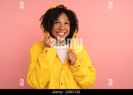Smiling dark skinned curly woman in yellow raincoat with hood has good mood, looking up at rain and blank space for advertising, promotional content, Stock Photo