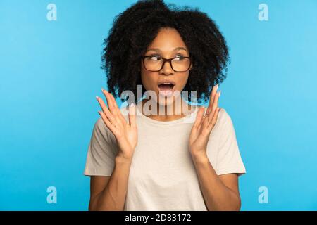 Surprised dark skinned woman in glasses with widely opened mouth, gesturing palms, looking aside at blank space for advertisement, shocked by news, di Stock Photo