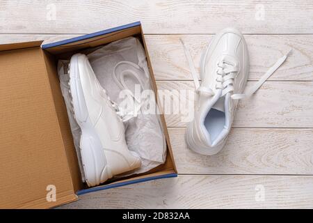 One white chunky sole sneakers in a brown cardboard box, the other on the white wood floor. Open box with new comfortable shoes for active lifestyle Stock Photo