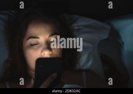 Young woman or girl lying in bed at night uses phone, reads news, writes messages or hangs out on social networks, copy space for text