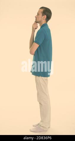 Full body shot profile view of young bearded man standing while thinking against white background Stock Photo