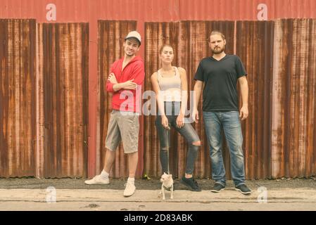 Happy group of friends smiling and standing while posing against old rusty sheet wall with woman holding cute dog Stock Photo