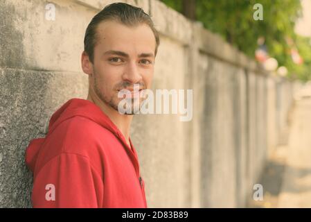 Young happy bearded man smiling while wearing red hoodie and leaning on old cement wall in the streets outdoors Stock Photo