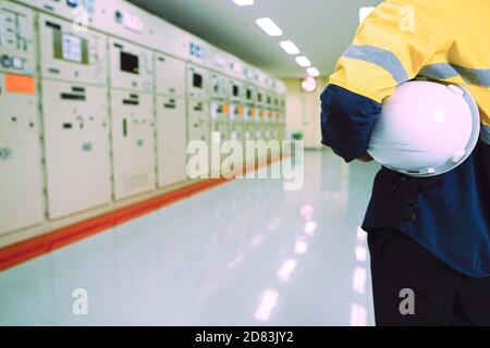Male engineer wearing a yellow uniform and wearing a white safety hat, inspecting electrical systems in a large power plant. Stock Photo