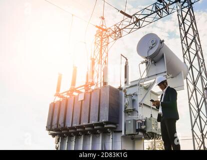 A low angle image of a businessman wearing a black suit, standing looking at a large power transformer with blue sky to be background, Concept about b Stock Photo