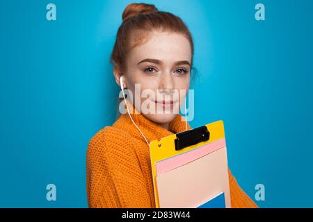 Close up photo of a ginger student with freckles posing with folders and earphones on a blue studio wall in a yellow sweater Stock Photo