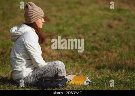 Brunette woman wearing hat sitting on the grass with eyes closed in the park on a beautiful sunny autumn day. Relaxing after hard day. Meditation Stock Photo
