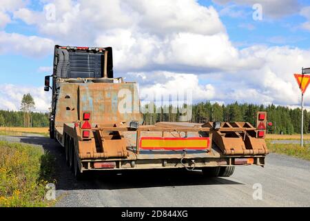 Semi truck pulling empty low loader trailer on a narrow country road making a right turn to the highway. Rear view, copy space top right. Stock Photo