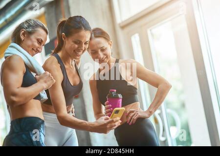 Group of three young beautiful and happy sportive women taking selfie on smartphone while resting after workout at gym, exercising together. Sport, wellness and healthy lifestyle Stock Photo