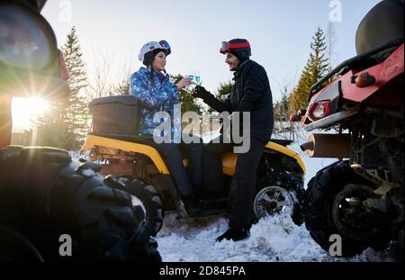 Young couple wearing winter ski suits and helmets, sitting on yellow ATV holding two glasses with blue champagne looking at each other. Sunny winter day. Huge wheels of other ATV are on foreground Stock Photo