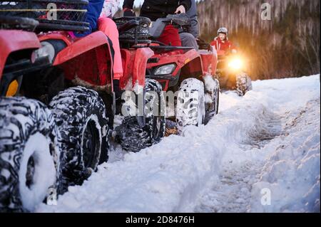 Close up of people riding red all-terrain vehicles down snowy hill. Quad riders driving quad bikes with black snowy wheels on snow-covered trail. Concept of winter activities and quad biking. Stock Photo