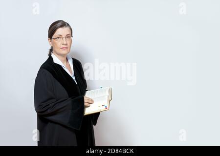 Middle aged female judge in black gown with a law book, neutral background, copy space. Stock Photo