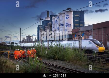 Network Rail track workers busy on the West Coast Main Line at Warrington Bank Quay station with the Unilever soaps factory in the early evening. Stock Photo