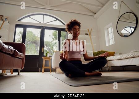 Low angle view of beautiful young woman sitting on fitness mat at home in lotus position while meditating Stock Photo