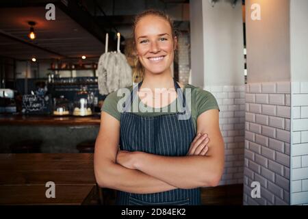 Portrait of beautiful young female waitress standing with arms crossed in cafe and wearing apron Stock Photo