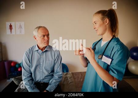 Young female therapist with stethoscope explaining exercise to senior patient with hand movement Stock Photo