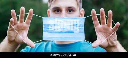 Scared Caucasian man holding blue medical mask in front of his face outside. Surgical disposable breathing mask as protection against coronavirus Stock Photo