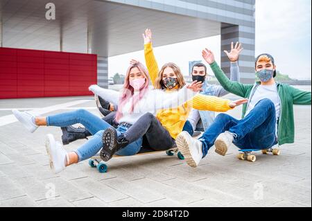 Cool teen friends in coronavirus time having fun, riding on skateboards in the city streets - Happy teenagers smiling under the  face masks - New norm Stock Photo