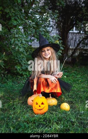 Funny child girl in witch costume for Halloween sitting on pumpkin Jack outdoor in park Stock Photo