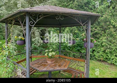 Hexagonal gazebo with ornate elements, with benches around, wooden table in middle, with vase of white flowers and wicker baskets with flowers around Stock Photo