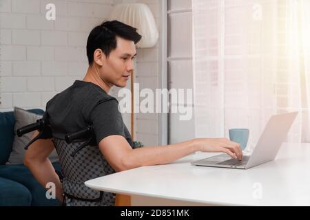 Happy disabled Asian man sitting in a wheelchair And working with computer at home, The concept of Technologies for the convenience of people with dis Stock Photo