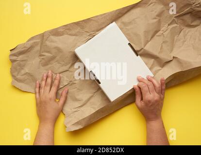 rectangular white cardboard box and two female hands are wrapped with brown paper on a yellow background, top view Stock Photo