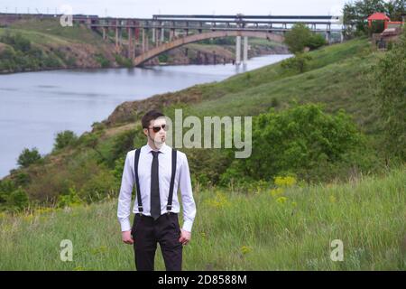 Portrait of stylish brunet young man, wearing white shirt, black tie, suspenders and glasses, walking on the meadow near river. Stock Photo