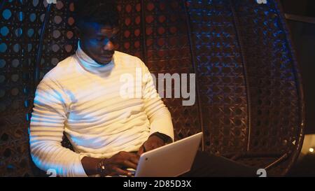 Young black man, freelancer sitting on the swing on the balcony and using laptop at night. High quality photo Stock Photo