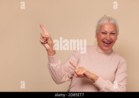 Amazed excited mature old woman pointing at copy space aside. Cheap price offer. Stock Photo