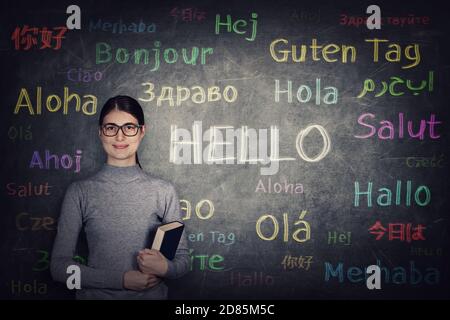 Young woman teacher or student, wearing eyeglasses and holding a book, stands in front of blackboard written with chalk the word hello in different la Stock Photo