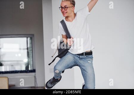 Happy hipster guy in casual clothes and with backpack jumping indoors against white wall Stock Photo