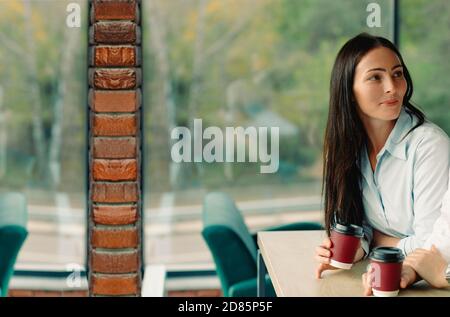 The brunette girl is resting and drinking coffee at the work coffee break. The concept of rest in a cafe during a break between work Stock Photo