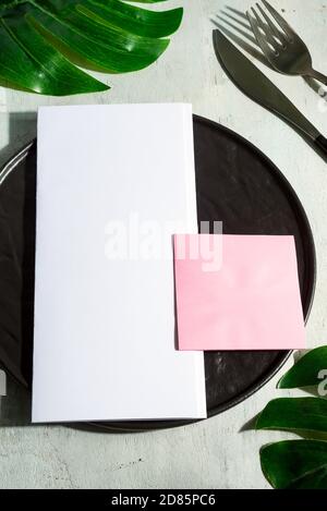 Served ceramic plate with knife fork and mock-up paper invitation cards on a light grey marble background decorated green leaves, copy space. Top view Stock Photo