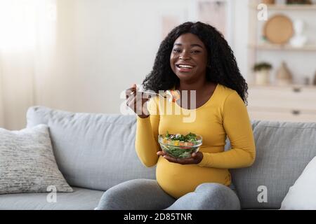 Happy pregnant black woman sitting on couch, eating fresh salad Stock Photo