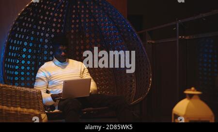 Young black man siting on the balcony swing and using laptop. Relaxation and entertainment at home. High quality photo Stock Photo