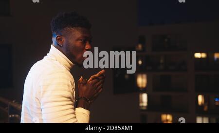 Young black man standing on balcony and rubbing his hands on cold night. High quality photo Stock Photo