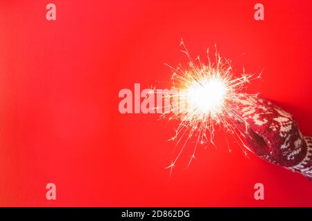 Hand in mitten holding burning sparkler on red background. Bengal fire on red-colored background. Sparks on burning sparkler in hand with knitted glov Stock Photo