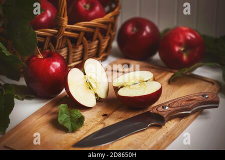 On the kitchen table still life - wicker basket with red ripe sweet apples, next to it a wooden board on which lies an Apple cut in half and a sharp k Stock Photo